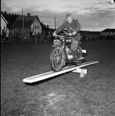 Arbogas Stjärnknutte
Balansövning med motorcykel på lutande bräda. Tävling.
Bilden är tagen på kvällen. I bakgrunden syns villor och publik.