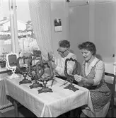 Börjesson tillverkar Arbogaspeglar
Ett par sitter vid köksbordet, i villakvarteret, med en uppsättning lika dana spegelramar uppställda. Kvinnan har förkläde på sig. På väggen hänger en almanacka.