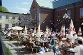 Arbogaträffen
En tillfällig servering på Rådhusets innegård. Folk sitter på träbänkar och dricker öl eller äter glass. Parasollen är nedfällda.