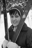 Gunvor Andersson deltar i skidtävlingen AT-loppet. Här står hon med sina längdskidor. Hon har mössa och lovikavantar på sig.
(AT kan betyda Arboga Tidning)