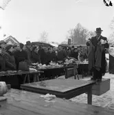 Auktion utomhus på Hamntorget. Auktionsförrättaren står på en trälåda. Han är iklädd rock och hatt. Han håller i en kavaj och ett par byxor. På ett uppbyggt bord står porslin och andra småsaker. Köparna är iklädda ytterkläder och en del av dem har paraply. Till vänster skymtar ett hamnmagasin och till höger Texacomacken.