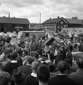 Auktion på Hamntorget. Auktionsförrättare med assistent som visar upp en stol. Publiken trängs. Bilar står parkerade på Hamntorget och längs Östra Hamngatan. I bakgrunden ses skylten till SWB Transport och ett hamnmagasin.