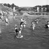 Gott om folk vid Villagatsbadet. Västerbron i bakgrunden. Badbryggor i Arbogaån och sittbänkar i sluttningen till vänster.