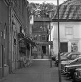 Västra delen av Stora torget med frisör och café Saga. Två flickor med barnvagnar går i Baggens gränd. Stadskällaren, på Nygatan, ses längst bort. Till höger i bild ses Duka-butiken och bilar som står parkerade.
