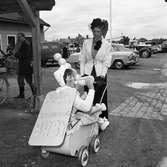 Barnens Dag firas. Här en mamma med en ovantligt stor baby i vagnen. Bakom dem skymtar diverse personbilar på parkeringen.