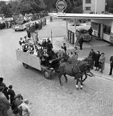 Barnens Dag firas. Två hästar drar en vagn med Barnens Dags Prins och Prinsessa; Erik-Gunnar Larsson och Margareta Lönnberg. Fler utklädda barn sitter på vagnen.
En lastbil kommer efter, i paraden. Till höger ses Shells bensinstation vid Jäderstullen. Publiken kantar Jädersvägen.