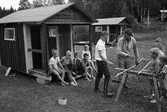 Barnens Dagskolonin. Flera barn (mest pojkar) utanför en liten campingstuga. Två vuxna män. Ytterligare stugor i bakgrunden.
Läs om Barnens Dags föreningen i Arboga Minnes årsbok 2002.