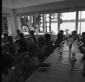 Barnens Dagskoloni vid Högsjön, väster om Arboga. Barnen sitter vid två långbord, i stugan, och äter mellanmål. De serveras ett glas mjölk och knäckebröd. Vacker utsikt, över sjön, genom de många fönstren. Det står en blomvas på bordet.