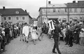Barnens Dag firas med en parad med utklädda barn. Först kommer figurer från Peter Pan.
Många människor har samlats för att titta på. Platsen är Stora torget.
I bakgrunden ses bland andra butiker Stures Möbler.