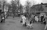 Barnens Dag firas på Sturevallen med karuseller och lottstånd. Människor i alla åldrar har kommit till festplatsen trots att det har regnat. I förgrunden ses en kvinna med ett barn i vagn. Hon har paraply med sig. I bakgrunden ses träden i Olof Ahllöfs park.
