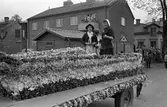 Barnens Dag firas med en kortege genom staden. Här är en blomsterprydd vagn med tre flickor på. Publiken står efter vägen och ser på.