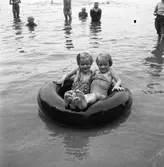 Barnleken. Två flickor, i baddräkt, flyter på en inneslang till ett däck. Andra badande ses i bakgrunden.
Inget årtal angivet.