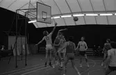 Herrlag spelar basket i plasthallen. Två barn sitter på en bänk och tittar på.