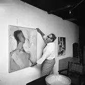 Bertil Levin ordnar konstutställning i Stallet, Västerlånggatan 1.
Man som hänger upp en tavla. Några möbler skymtar.