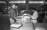 Två flickor vid bibliotekets lånedisk, de är Anita Blomander och Lena Johansson. Bakom disken sitter Elisabeth Källberg. I bakgrunden ses bokhyllor fyllda med böcker. Bakom Elisabeth står ett bord med en telefon på.
Bilden publicerades i Västmanlands Läns Tidning 14 mars 1972.