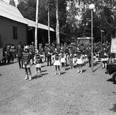 Blåsorkester med ungdomar, klädda i uniform.
Platsen ser ut att vara Folkets park. En buss är parkerad.