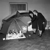 Utställning på Folkan/Medborgarhuset om olika dukningar. Här är det dukat för en utflykt till stranden med solparasoll, kaffepanna, termos, en korg med frukt, en flaska mjölk, bröd på en skärbräda och två matlådor av aluminium.
Två kvinnor ser på utställningen. På väggen, bakom parasollet, hänger en tavla.