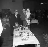 På Folkan/Medborgarhuset visas olika dukningar. En familj besöker utställningen och har stannat vid ett bord med dessert. Frukter som banan och apelsin ingår i efterrätten. Kanske är det glass på faten.
Kvinnan bär päls och hatt med flor. Mannen har kostym. Han bär minstingen på armen. Storebror står vid mamma.
Andra utställningsbesökare ses i bakgrunden.