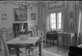 Bostadsinteriör, Crugska gården. Dekorerade väggar. Ett flertal olika möbler bland annat ett bord och fyra stolar. Till höger anas ett piano. En krukväxt på ett bord vid fönstret. Tavlor på väggarna. En ljuskrona i taket.