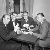 Distriktsmästerskap i bridge
Fyra män sitter vid ett bord och spelar kort. I fönstret står en ljusstake.
Bridge-DM