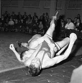 Brottning. Arboga AK mot Sala. Brottare på mattan. I publiken sitter en man med kamera.
AAK, Arboga AtletKlubb