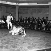 Brottning. Arboga AK mot Sala. Domare och två tävlande på mattan. Publiken sitter efter väggarna.
Arboga AtletKlubb