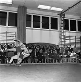 Tävlingen Fyrstadsbrottning mellan Arboga Atletklubb, Eslöv, Uddevalla och Falkenberg.
Två män ovanför brottarmattan. Publik i gymnastiksalen.