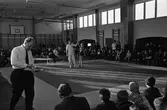 Brottning för ungdomar. Matcher pågår på två mattor samtidigt. Närmast kameran har en segrare korats. Domaren håller upp brottarens arm. Publiken sitter efter väggarna i gymnastiksalen.
1960-talet