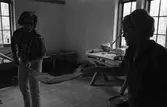 Brödbakning vid Jäders bruk. Två kvinnor i en bakstuga/bagarbod. En kvinna håller i en brödspade. Möjligen bakar de knäckebröd. Vid det ena fönstret finns ett bakbord med brödkavel, mjölpåse, bunke och brödkavel.