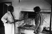 Brödbakning vid Jäders bruk. Två kvinnor i en bakstuga/bagarbod. En kvinna håller i en brödspade som hon sticker ini ugnen. De bakar knäckebröd.