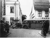 En buss, från Uppsala, stannar utanför Öhrman & Melander på Nygatan. En färgad man kliver av.
Människor på trottoarerna. Cyklar och en bil.