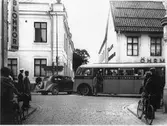 En buss, från Uppsala, stannar utanför Öhrman & Melander på Nygatan. En färgad man kliver av.
Människor på trottoarerna. Cyklar och en bil.