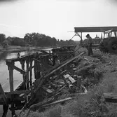 Hamnen. En del av kajen rivs. Män i arbete med att plocka ner bräder. En traktor står nära kantena av Arbogaån. Det långa röret, en bit ovanför marken, tillhör Lantmännens Silo. I bakgrunden ses Arboga Kvarn och Maltfabrik samt tornet på Heliga Trefaldighets kyrka.