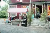 ICA-butiken Lövsta Handel. En man är på väg ut med en matkasse i varje hand. Trappan är lövad. En liten hund är bunden vid en av björkarna. En pojke står intill ett torgstånd där diverse campingutrustning säljs. Vid cykelstället är en liten, svart hund bunden. Affärens skyltfönster är täckta av reklamaffischer. Det är sommar.