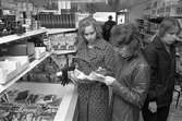 Två elever studerar priser i frysdisken på mataffären Ringköp. Den vänstra flickan är Eva Sörensen. Andra kunder skymtar i affären.