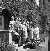 Elever vid Fellingsbro folkhögskola
Nio unga kvinnor och två män står på en trappa utanför en port. Väggen är täckt av vildvin. De bär på en karmstol. Två resväskor och annat bagage står på grusgången. En man tittar i en anteckningsbok.