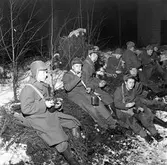 FBU-ugdomar övar i skogen. Det är vinter och snö. Grabbarna är klädda i yllerockar och mössor med öronlappar. De sitter på granris. De äter ur sina plåtburkar.
FBU betyder Frivillig Befälsutbildning