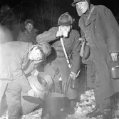 FBU-ungdomar övar
Unga män, iklädda ylleöverrockar och öronlappsmössor. De står i snön och lagar varm dryck. En dricker. De har sina matburkar (