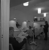 Frisör Axel Fritiof Strandberg (kallad Atschy) i salongen på det nyöppnade ålderdomshemmet Strömsborg.
Axel klipper en kund. En kvinna sitter i torkhuven. En man läser en tidning.