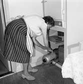 Fru Blomberg i köket. Hon tar fram dammsugaren ur skåpet under kylskåpet. Där står också några kakburkar. Kvinnan har förkläde på sig.
Hon är gift med Jakob Blomberg som arbetar på Arboga Mekaniska Verkstad.
AMV
