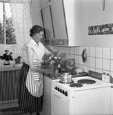 Fru Blomberg, (maka till Jakob Blomberg på Arboga Mekaniska Verkstad) i köket. Hon står vid diskbänken och arrangerar blommor i en vas. Kaffepannan står på spisen. Det står krukväxter på fönsterbrädan.
AMV