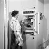 Fru Blomberg, hustru till Jakob Blomberg, på Arboga Mekaniska Verkstad, visar sitt kylskåp. I kylskåpet ses några skålar och en kakburk. En kökssoffa anas till höger.
Kvinna, iklädd förkläde, står intill ett kylskåp.
AMV