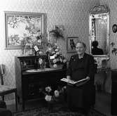 Fru Karlsson, 70 år
Kvinna som sitter i sitt hem. Det står blomsterbuketter, i vaser, på sekretären. På väggen hänger en stor spegel, en tavla samt ett fotografi av en hund.