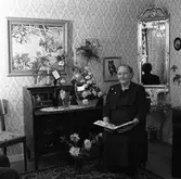 Fru Karlsson, 70 år
Kvinna som sitter i sitt hem. Det står blomsterbuketter, i vaser, på sekretären. På väggen hänger en stor spegel, en tavla samt ett fotografi av en hund.