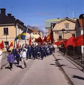 Första maj. Demonstrationståg med röda fanor och plakat.
Först går ett par med en svensk flagga. Sedan följer Arboga Blåsorkester under ledning av Håkan Harrysson.
I bakgrunden ses kvarteret Stadsgården till vänster och Kungsgården till höger.
Kapellbron är dekorerad med röda fanor.
Socialdemokrater demonstrerar.