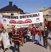 Första maj. Demonstrationståg på Kapellbron. Berit Oscarsson längst till vänster. 
Människor marscherar med banderoller.
I bakgrunden ses kvarteret Stadsgården till vänster och Kungsgården till höger.
Socialdemokrater