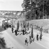 Första maj. Demonstration. Fanbärarna går först. Sedan följer människor med plakat. De har tågat över Västerbron och är på väg mot Folkets Park. Till höger ligger Törnberget.
I bakgrunden ses Villagatsbadet och bebyggelsen på Nästkvarnsområdet
Socialdemokrater