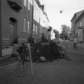 Gatuarbete på Kapellgatan. Två män arbetar i gropen. De har ställt upp bockar runt hålet. Några barn tittar på.