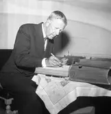 Konung Gustaf Vl Adolf är på Eriksgata. Han har just sett silversamlingen i Heliga Trefaldighetskyrkan. Här skriver han i kyrkans gästbok.
För värdskapet, under kungens besök, svarade stadsfullmäktiges ordförande Jonas Carlsson och kommunalborgmästare Danliel Ekelund. Den organisationskommitté som ansvarade för arrangemangen, hade hos drätselkammaren begärt en summa av 3000 kronor för att täcka kostnaderna vid kungabesöket. Beloppet beviljades. 
Tiden för kungens besök var beräknad till 130 minuter.