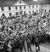 En stor publik har samlats på Stora torget för att se och höra konungen. Gustaf Vl Adolf gästar Arboga under sin Eriksgata. Framför Stadsgården står en stor skara människor, längst fram står männen i hemvärnet.
För värdskapet, under kungens besök, svarade stadsfullmäktiges ordförande Jonas Carlsson och kommunalborgmästare Danliel Ekelund. Den organisationskommitté som ansvarade för arrangemangen, hade hos drätselkammaren begärt en summa av 3000 kronor för att täcka kostnaderna vid kungabesöket. Beloppet beviljades. 
Tiden för kungens besök var beräknad till 130 minuter.
